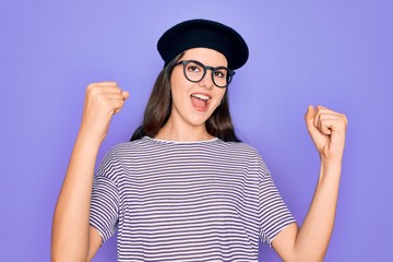 Young beautiful brunette woman wearing glasses and french beret over purple background celebrating surprised and amazed for success with arms raised and open eyes. Winner concept.