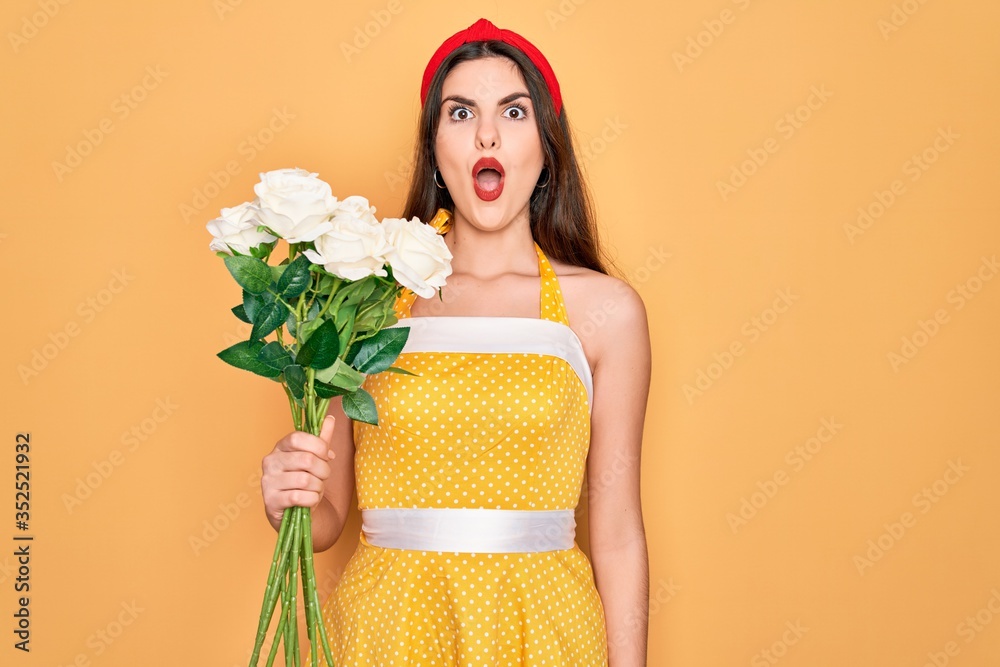 Poster Young beautiful pin up woman wearing 50s fashion vintage dress holding boquet of roses flowers scared in shock with a surprise face, afraid and excited with fear expression