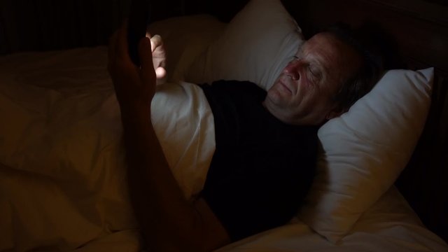 Man using a smartphone lying on bed before sleep at night in home, close up. The light from the phone is illuminating his face