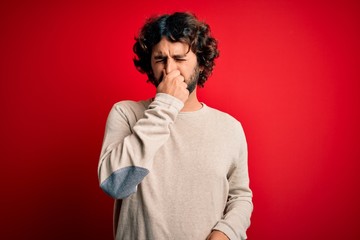 Fototapeta na wymiar Young handsome man with beard wearing casual sweater standing over red background smelling something stinky and disgusting, intolerable smell, holding breath with fingers on nose. Bad smell