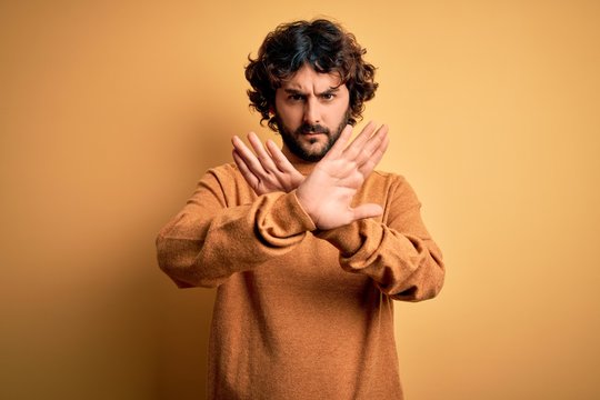 Young handsome man with beard wearing casual sweater standing over yellow background Rejection expression crossing arms doing negative sign, angry face