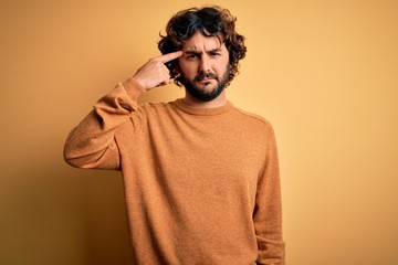 Young handsome man with beard wearing casual sweater standing over yellow background pointing unhappy to pimple on forehead, ugly infection of blackhead. Acne and skin problem