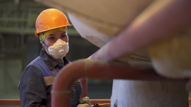Portrait of a women worker at the factory. Stock footage. Female wearing protective face mask and an orange helmet while standing and controlling working process near machine at the industrial plant.