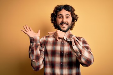 Young handsome man with beard wearing casual shirt standing over yellow background amazed and smiling to the camera while presenting with hand and pointing with finger.