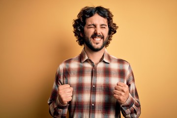 Obraz premium Young handsome man with beard wearing casual shirt standing over yellow background excited for success with arms raised and eyes closed celebrating victory smiling. Winner concept.