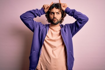 Young handsome sporty man with beard wearing casual sweatshirt over pink background doing funny gesture with finger over head as bull horns