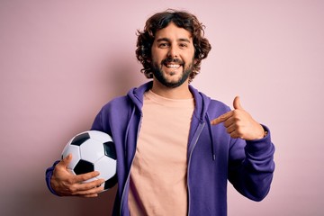 Handsome player man with beard playing soccer holding football ball over pink background with...
