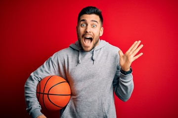 Young sports man holding basketball ball over red isolated background very happy and excited, winner expression celebrating victory screaming with big smile and raised hands