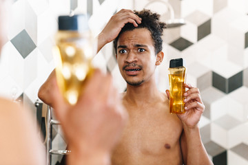 Photo of african american man holding shampoo and looking at mirror