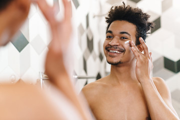 Photo of african american man applying face cream and looking at mirror