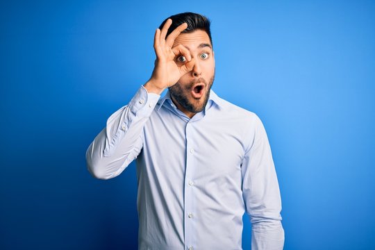Young handsome man wearing elegant shirt standing over isolated blue background doing ok gesture shocked with surprised face, eye looking through fingers. Unbelieving expression.