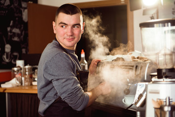 Professional barista holding metal jug warming milk using the coffee machine. Happy young man preparing coffee at counter