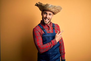 Young rural farmer man wearing bib overall and countryside hat over yellow background cheerful with a smile of face pointing with hand and finger up to the side with happy and natural expression