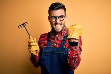 Young gardener man wearing working apron using gloves and tool over yellow background annoyed and...