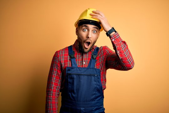 Young builder man wearing construction uniform and safety helmet over yellow isolated background surprised with hand on head for mistake, remember error. Forgot, bad memory concept.