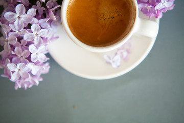 Fresh espresso in a white cup and lilac flowers on the background.