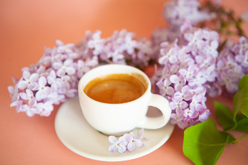 Fresh espresso in a white cup and lilac flowers on the background.