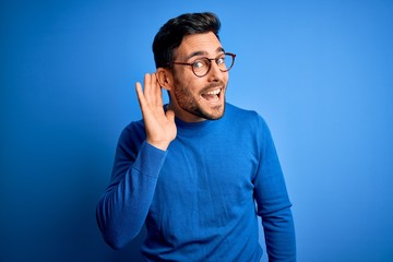 Young handsome man with beard wearing casual sweater and glasses over blue background smiling with...