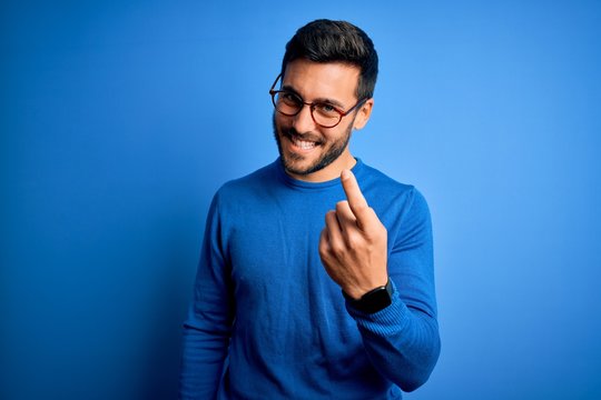 Young handsome man with beard wearing casual sweater and glasses over blue background Beckoning come here gesture with hand inviting welcoming happy and smiling