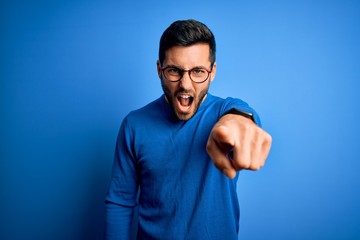 Young handsome man with beard wearing casual sweater and glasses over blue background pointing...