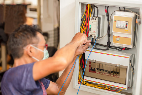 Electrician With Medical Mask Engineer Works With Electric Cable Wires Of Fuse Switch Box.