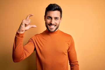 Young handsome man with beard wearing casual sweater standing over yellow background smiling and confident gesturing with hand doing small size sign with fingers looking and the camera. Measure