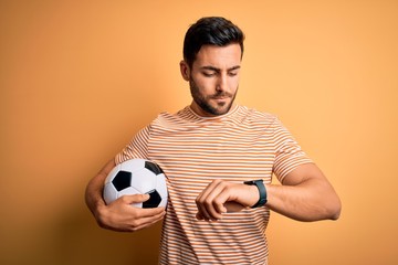 Handsome player man with beard playing soccer holding footballl ball over yellow background Checking the time on wrist watch, relaxed and confident