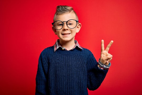 Young little caucasian kid with blue eyes standing wearing smart glasses over red background smiling with happy face winking at the camera doing victory sign. Number two.