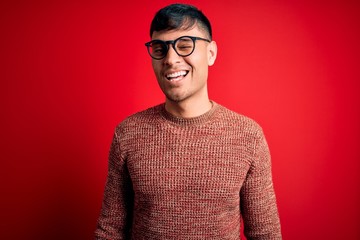 Young handsome hispanic man wearing nerd glasses over red background winking looking at the camera...