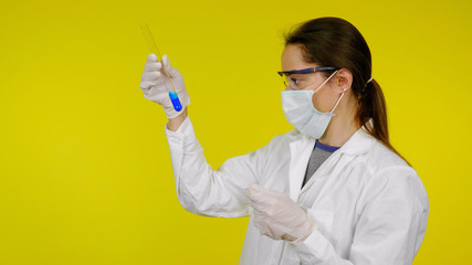 Doctor in a medical mask, goggles and latex gloves looks at the tests in the tube. Young girl in a white coat on yellow background holds hand a tube with blue liquid