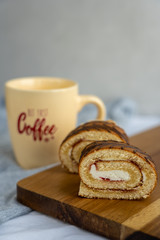 Two slices of sponge roll are laying on wooden cutting board against background of coffee cup