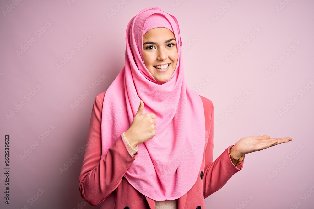 Canvas Prints Young beautiful girl wearing muslim hijab standing over isolated pink background Showing palm hand and doing ok gesture with thumbs up, smiling happy and cheerful