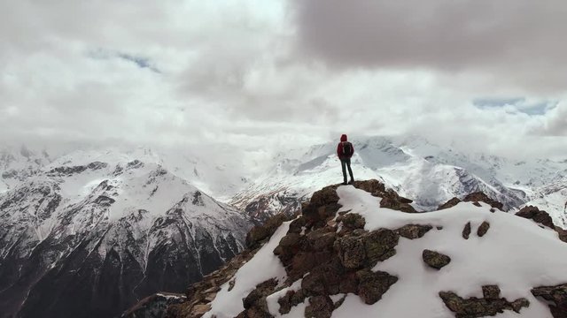 A male traveller with a backpack stands on top of a cliff against the backdrop of snow-capped mountain peaks and low clouds. Aerial view. Flying the camera past a man in a storon cliff