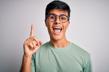 Young handsome man wearing casual t-shirt and glasses over isolated white background pointing finger up with successful idea. Exited and happy. Number one.