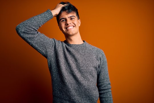 Young handsome man wearing casual sweater standing over isolated orange background smiling confident touching hair with hand up gesture, posing attractive and fashionable