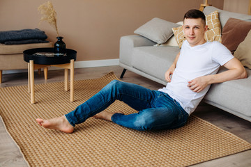 A young guy at home sitting on the floor in a beige interior barefoot and looking at the camera. Stay indoors.