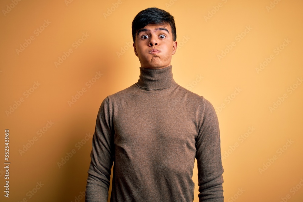 Poster Young handsome man wearing casual turtleneck sweater over isolated yellow background puffing cheeks with funny face. Mouth inflated with air, crazy expression.