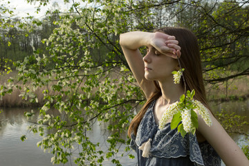A young girl with long blonde hair, in a blue dress, raised her hand to her face, closed her eyes, she is stuffy and hot in the heat. the girl has in her hands a flowering branch of bird cherry.