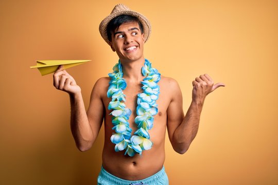 Young tourist man on vacation wearing swimwear and hawaiian lei flowers holding paper plane pointing and showing with thumb up to the side with happy face smiling