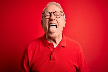 Grey haired senior man wearing glasses and casual t-shirt over red background sticking tongue out happy with funny expression. Emotion concept.