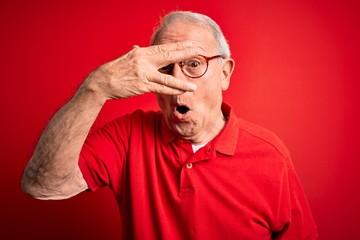 Obraz premium Grey haired senior man wearing glasses and casual t-shirt over red background peeking in shock covering face and eyes with hand, looking through fingers with embarrassed expression.