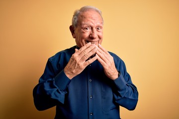 Grey haired senior man wearing casual blue shirt standing over yellow background laughing and...
