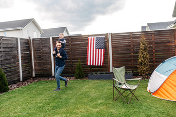 Backyard games. A young father is running and carrying his baby son on the shoulders, they are...