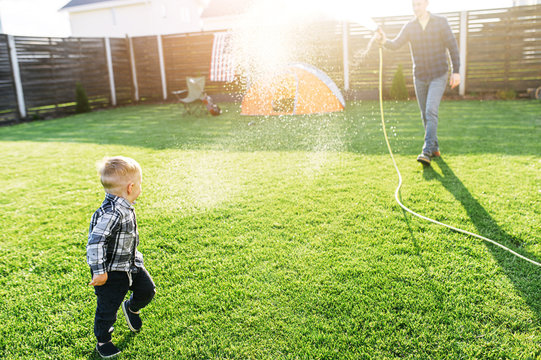 Young father and little cute son spend active time together in the backyard. Dad is watering son from hose, a boy is running away and laughing