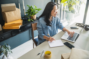 Young business woman working online e-commerce shopping at her shop. Young woman seller prepare parcel box of product for deliver to customer. Online selling, e-commerce.