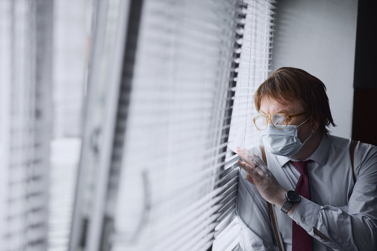 Aged empolyee in hygienic mask looking on street pulling back blinds on window
