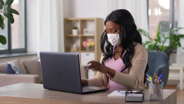 quarantine, remote job and pandemic concept - african american woman wearing face protective medical mask for protection from virus disease with laptop computer working at home office