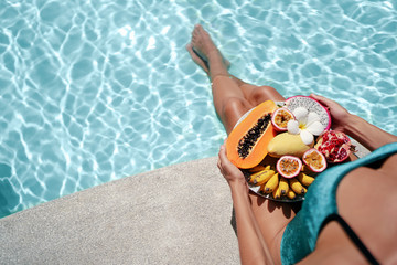 Enjoying vacation. Young woman in swimwear with tropical fruits plate sitting near swimming pool.