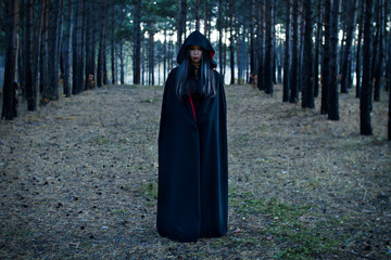 Vampire Woman in a gloomy forest. Beauty Sexy Vampire Girl in a cloak with a hood. Sexy Witch. Wicked look and pale skin. Attractive model girl in Halloween costume and make up.