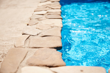 The pool with clear blue water is ideal for relaxing at home. The deep pool is lined with decorative stone along the edge, the sun is reflected in the water.